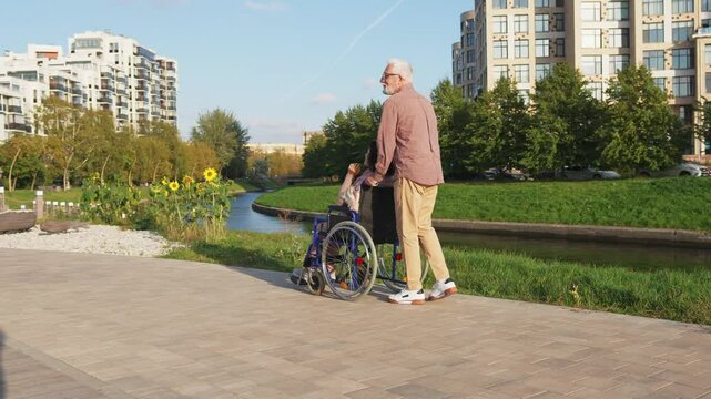 Senior woman in wheelchair walking with caregiver old man on road. Family couple man husband supporting taking care of paralyzed woman wife in chair for people with disability outdoor. Rehabilitation