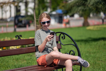 Fototapeta premium Young girl with long blonde hair sitting on bench in park, laughing and talking on smartphone