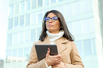 Businesswoman stands in front of an office building, holding a digital tablet and contemplating her...