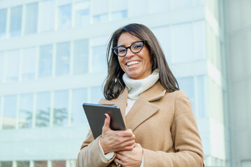 Fototapeta premium Businesswoman is seen outside an office building, happily holding a tablet and dressed in a camel coat and glasses