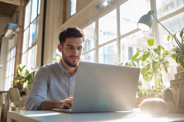 High-resolution brightly lit photorealistic candid photograph of a young marketer focused on his laptop in a bright, airy office with a joyful ambiance. The photograph is styled like a high-end
