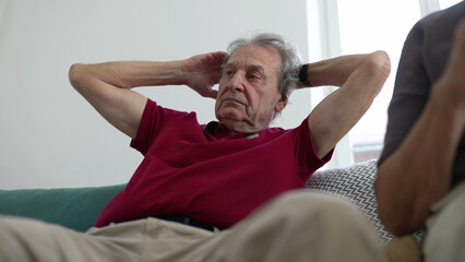 Obraz premium Elderly man in a red shirt relaxing on a couch with hands behind his head, contemplating, indoors, showcasing a moment of relaxation and comfort in a home environment, senior lifestyle