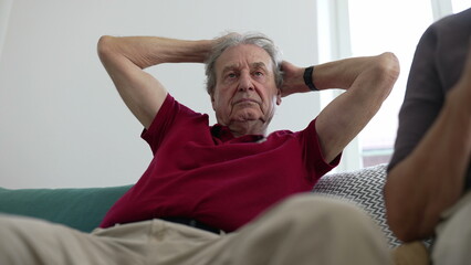 Elderly man in a red shirt relaxing on a couch with hands behind his head, contemplating, indoors,...