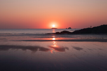 Obraz premium Sunset over the ocean sea and beach with silhouette of rocks, one adult person and reflections, pink and blue, Cornwall UK
