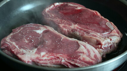 Close-up of raw steaks in a pan, starting to cook, lightly salted, fresh beef ready for cooking, sizzling sound, home kitchen, culinary preparation, delicious meal in progress, detailed shot