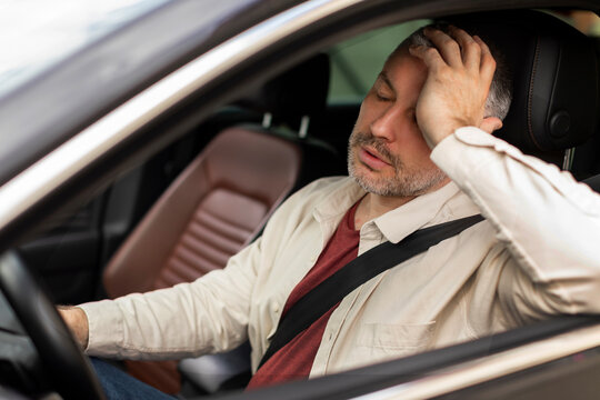 Sad middle aged man driving car, feeling tired and touching head, holding steering wheel in auto