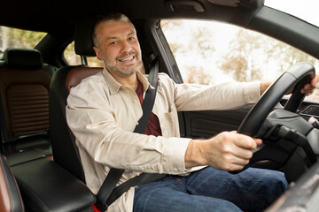 Positive sitting inside his own luxury car, holding hand on steering wheel and smiling at camera. Transportation, automobile renting