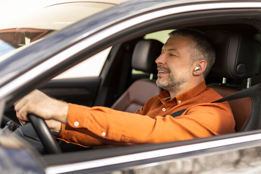 Middle aged businessman driver with wireless earphones or hands free device driving car, holding steering wheel and smiling