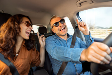 Joyful spouses in sunglasses enjoy driving luxury car, dancing to music and singing, going on vacation on weekend, windshield view