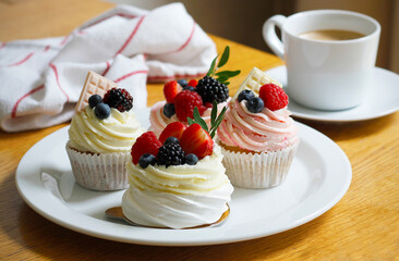 Cupcakes and mini pavlova cakes decorated with fresh berries and white chocolate on a large white plate, a cup of coffee and a cloth napkin in the background. Selective focus.