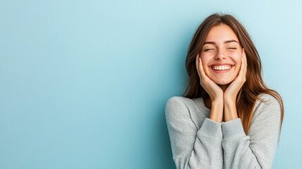 Young woman smiling joyfully with her hands on her cheeks, expressing delight and happiness against a soft blue background.
