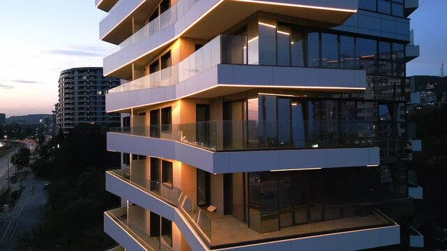 Aerial drone shot of a modern building facade illuminated by sleek LED lighting at dusk, capturing the contrast between the urban night skyline and contemporary architecture - Powered by Adobe