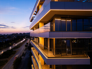 Aerial drone shot of a modern building facade illuminated by sleek LED lighting at dusk, capturing the contrast between the urban night skyline and contemporary architecture