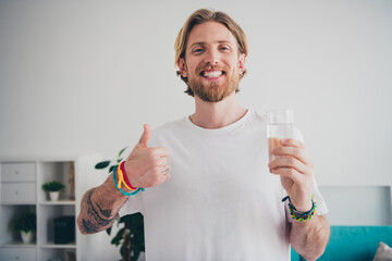 Photo of handsome young guy drink water glass thumb up dressed nightwear spend pastime spacious...