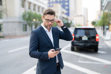 A handsome young businessman using his phone and standing on the street. Handsome smiling young businessman smartly dressed standing outdoors at the city street, using mobile phone