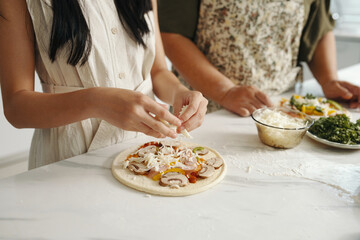 Close-up of girl putting vegetables on dough to make pizza together with her grandmother
