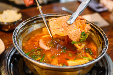 Tongs Holding a Large Piece of Pork from Kimchi Stew in a Metal Pot, Just After Being Cut with Scissors
