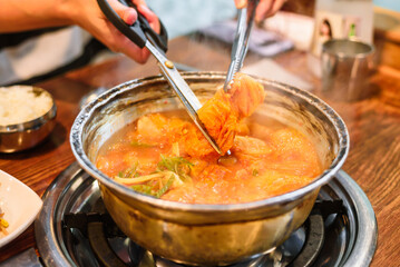 Tongs Holding a Long Piece of Kimchi from Kimchi Stew in a Metal Pot, Being Cut with Scissors
