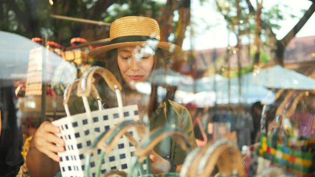 View Through The Window: Pleased Girl in a Linen Green Shirt and Straw Hat Picks Out Handmade Bag at the Weekend Jing Jai Market, Having Fun. Vacation, Asian Culture, Shopping Concept