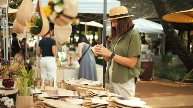 Pleased Girl in a Linen Green Shirt and Straw Hat Picks Out Handmade Plates at the Weekend Jing Jai Market, Having Fun. Vacation, Asian Culture, Shopping Concept