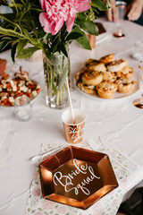 Decorations for a bachelorette party. On the table is a plate with the words ＂Bride Squad＂. In the background you can see food and flowers. The photo is bright and aesthetic.