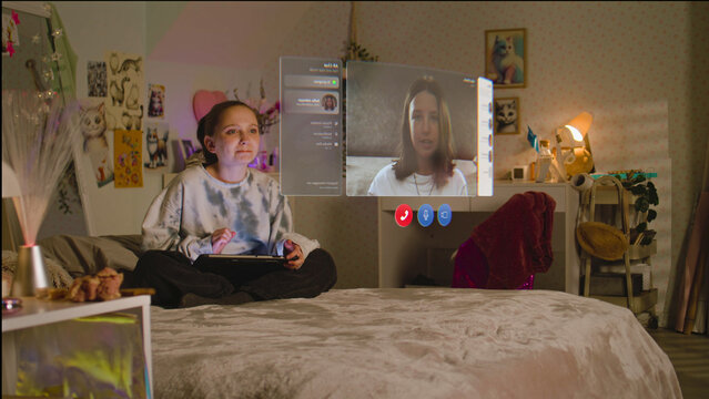 Teenager girl uses tablet computer, talks to friend in AR chat app using virtual reality, sits on bed at home. VFX graphics of holographic video call interface. Futuristic metaverse. Augmented reality