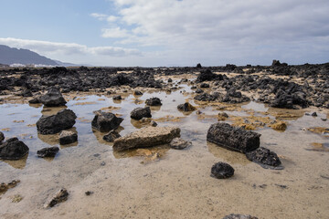White sand beach with black rocks of volcanic origin. Mountains in the background and sky with big white clouds. Caletón Blanco, Lanzarote, Canary Islands, Spain