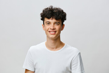 Young man with curly hair smiling in a plain white t shirt against a light gray background,...