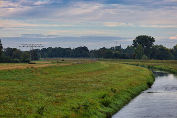 Obraz premium Blick auf den geschwungenen Verlauf der Neuen Luppe am Luppedamm, grüne Flusslandschaft mit Fernleitungsmast unter weitem Himmel, Leipzig, Sachsen, Deutschland
