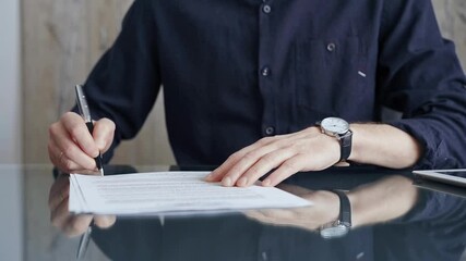 Businessman in dark blue t-shirt is signing contract at desk. Close-up of a male executive signing a legal document on a glass desk. Business people concept