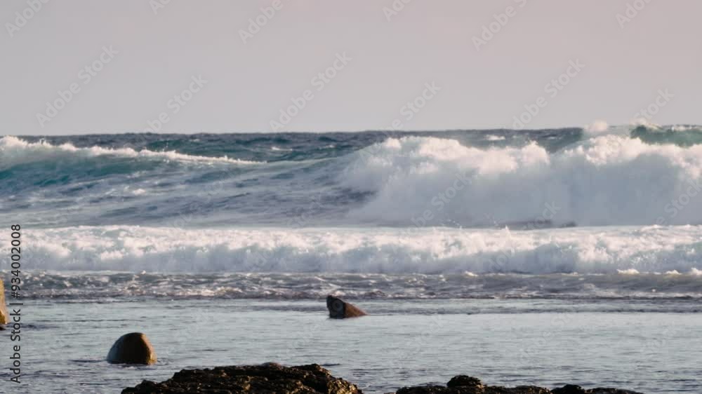 Poster waves roll over rocks at a coastal area under a colorful sky during sunset, creating a serene and dy