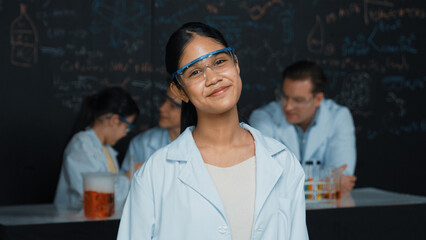 Girl looking at camera with arm folded while people doing experiment at laboratory. Cute student...