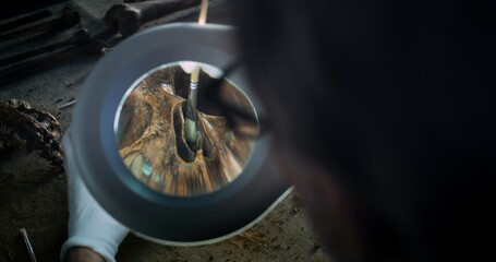 Mature paleoanthropologist or archaeologist cleans skull of ancient prehistoric human with tools, looks through magnifying lamp. Male scientist works with fossil remains in modern archaeological lab.