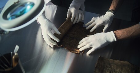 Two archaeologists in gloves carefully roll out ancient Egyptian papyrus on the table in...