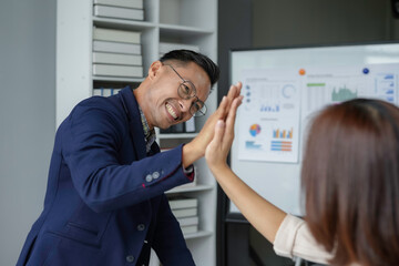 Two asian business people celebrating success giving high five in the office with charts and graphs in the background