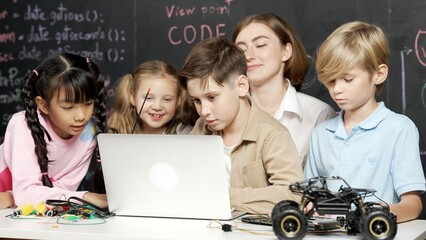 Closeup of boy using laptop programing engineering code and writing program while group of diverse kid holding controller in STEM technology classroom at blackboard written with prompt. Erudition.