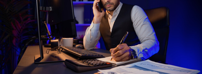 Smart businessman calling partner or investor on smartphone on panoramic view, writing data on paper work report to analyze sales channel discipline's product service at neon dark light room. Surmise.