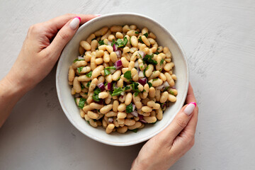 Female hands holding a bowl of homemade White Bean Salad with Herbs and Onion, top view. Flat lay, overhead, from above.