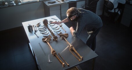 Archaeological laboratory: High angle of mature archaeologist in AR headset studying remains of prehistoric human skeleton. Scientist conducts historical research using virtual reality technology.