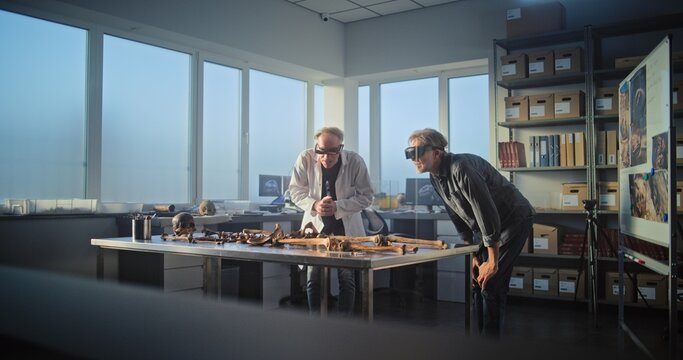 Two archaeologists wearing AR headsets study remains of prehistoric extinct human skeleton. Scientists conduct historical research using virtual reality technology, work in archaeological laboratory.