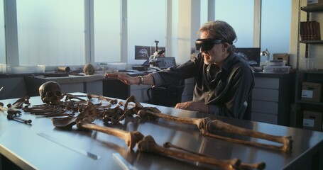 Mature archaeologist in VR headset studies remains of ancient human skeleton using augmented reality technology, conducts scientific research in archaeological lab illuminated by the sunset light.