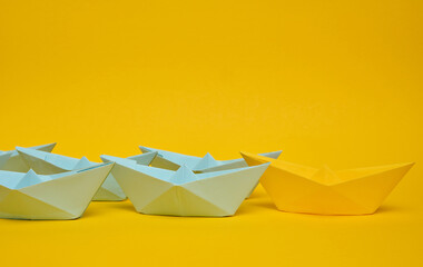 Group of blue paper boats follow yellow against a yellow background. Strong leader concept, mass...