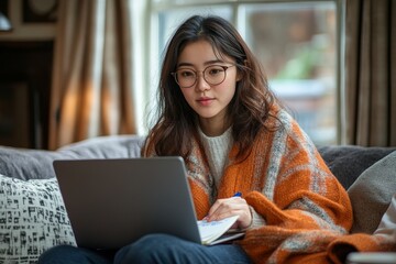 Asian woman wearing glasses writes notes on her notepad while working from home sitting in front of her laptop computer on a sofa in the living room of a house in Edinburgh, Scotland, Generative AI