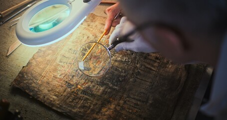 Senior archaeologist looks through magnifying lamp, studies ancient Egyptian papyrus with colleague...