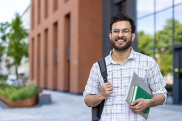 Portrait of a young smiling Indian male student standing on a university campus outside, holding a backpack and books in his hands, looking confidently at the camera