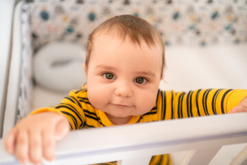 Cute baby boy in a yellow striped outfit trying to stand in his crib. Adorable toddler smiling and...