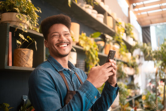 Smiling florist enthusiastically using smartphone in a lively flower shop. Happy flower shop worker in a brown apron checking messages surrounded by colorful plants and blossoms.