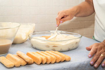 A hand spreads creamy mascarpone over soaked ladyfingers in a glass dish, finalizing a step in making tiramisu, an Italian dessert.