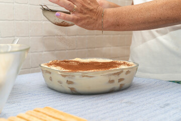 A person sifting cocoa powder over a layered tiramisu in a glass dish, showcasing the final step in...