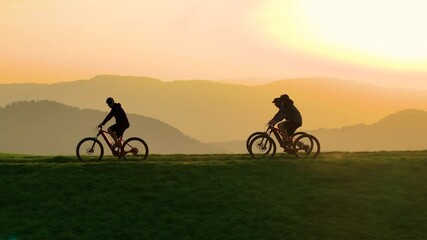 AERIAL, SILHOUETTE, LENS FLARE: Cyclists riding along a scenic hilltop at sunset. Vibrant glowing orange sky and the sheer beauty of an evening outdoor adventure in a picturesque country setting.